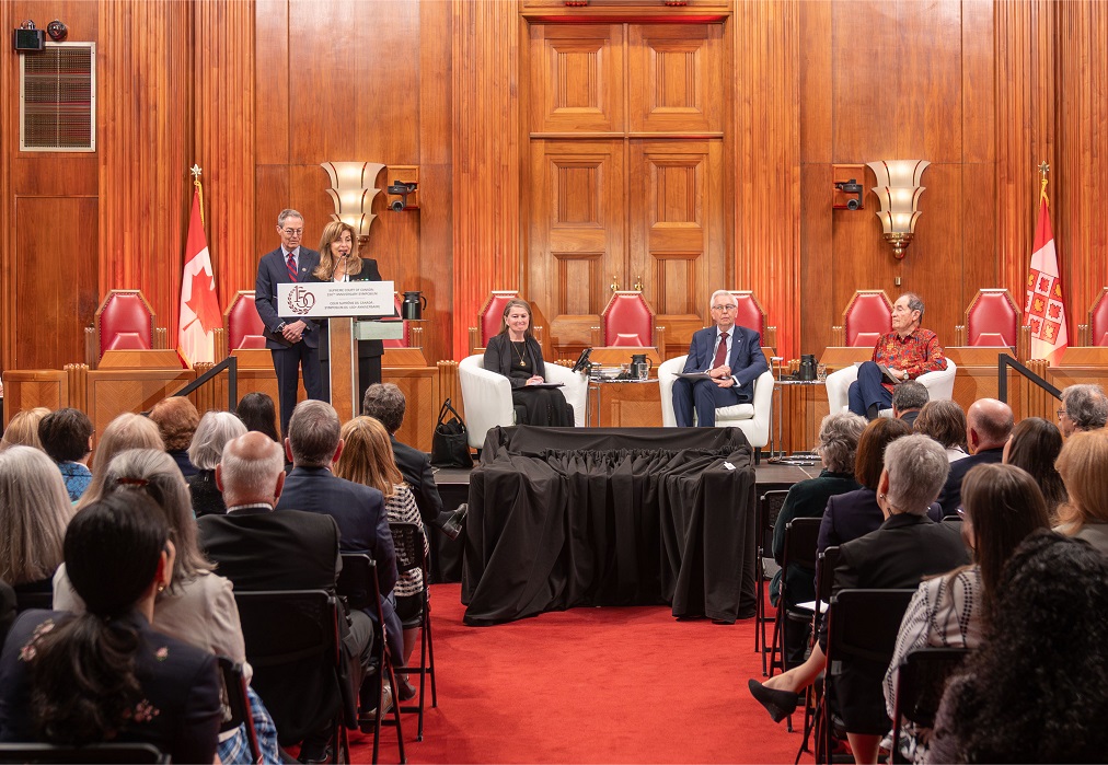 Justices Karakatsanis and Kasirer speak at a podium in the main courtroom, while panellists watch on from the stage