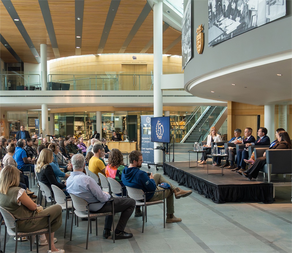 The judges sit on stage in front of an audience in a large atrium 