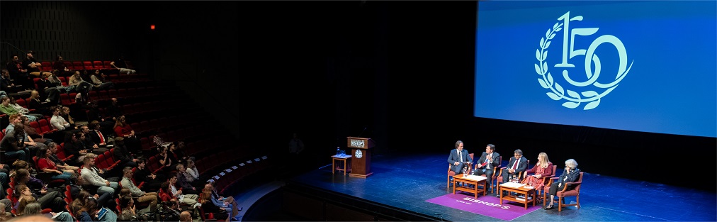 The judges sit on a stage in front of an audience in a large auditorium